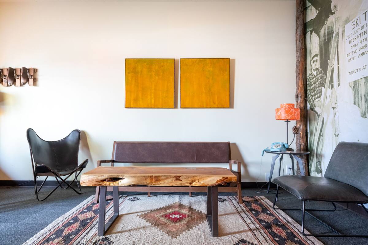 Una habitación con un banco de madera, dos sillas negras, un tapete estampado y una pared con dos cuadros amarillos.