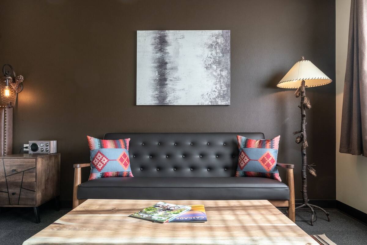 Una sala de estar moderna con un sofá oscuro, almohadones coloridos, una mesa de centro con una revista, una obra de arte en la pared, una lámpara y una mesita de noche con un radio.