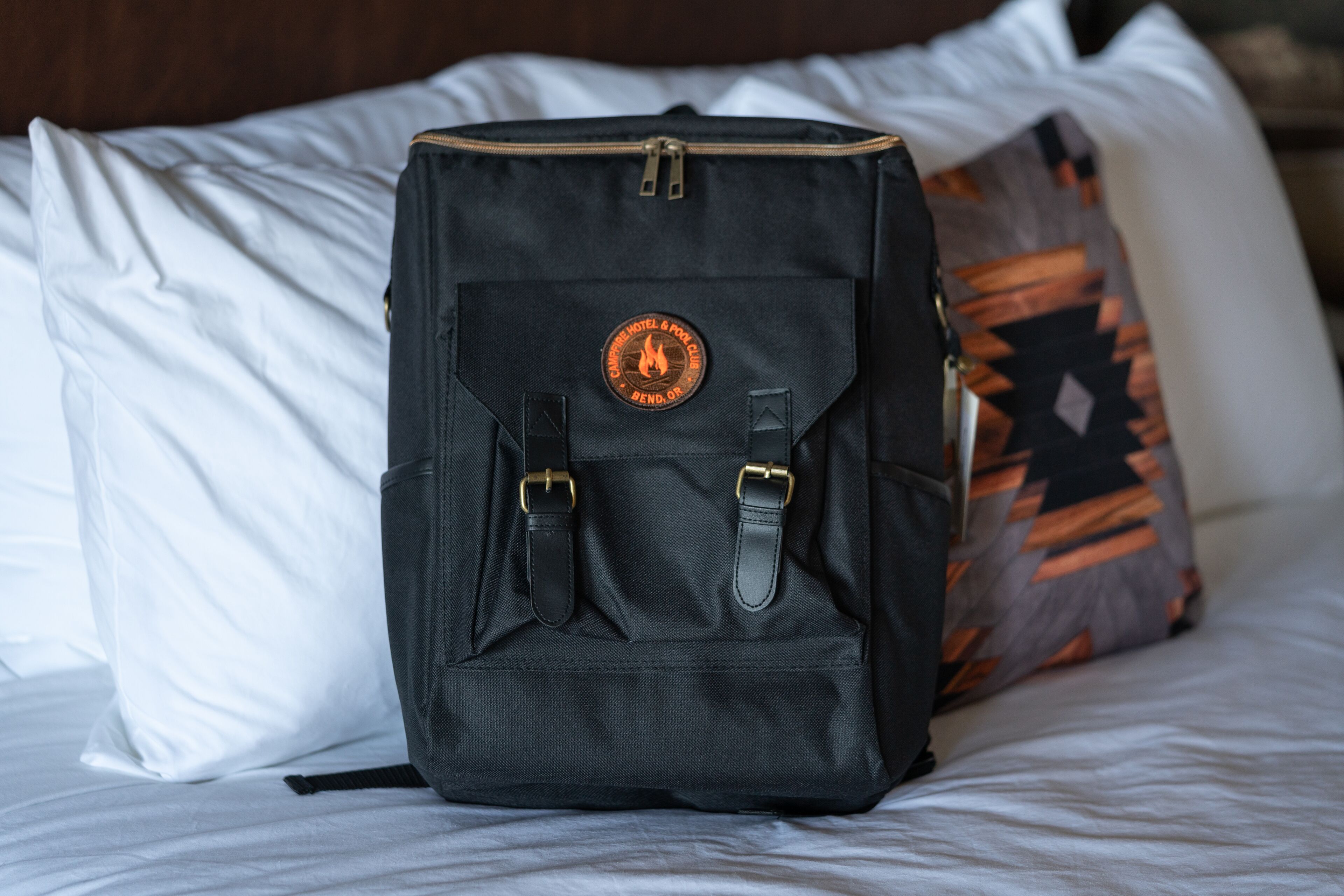 A black backpack with a logo on the front, placed on a bed with white and patterned pillows.
