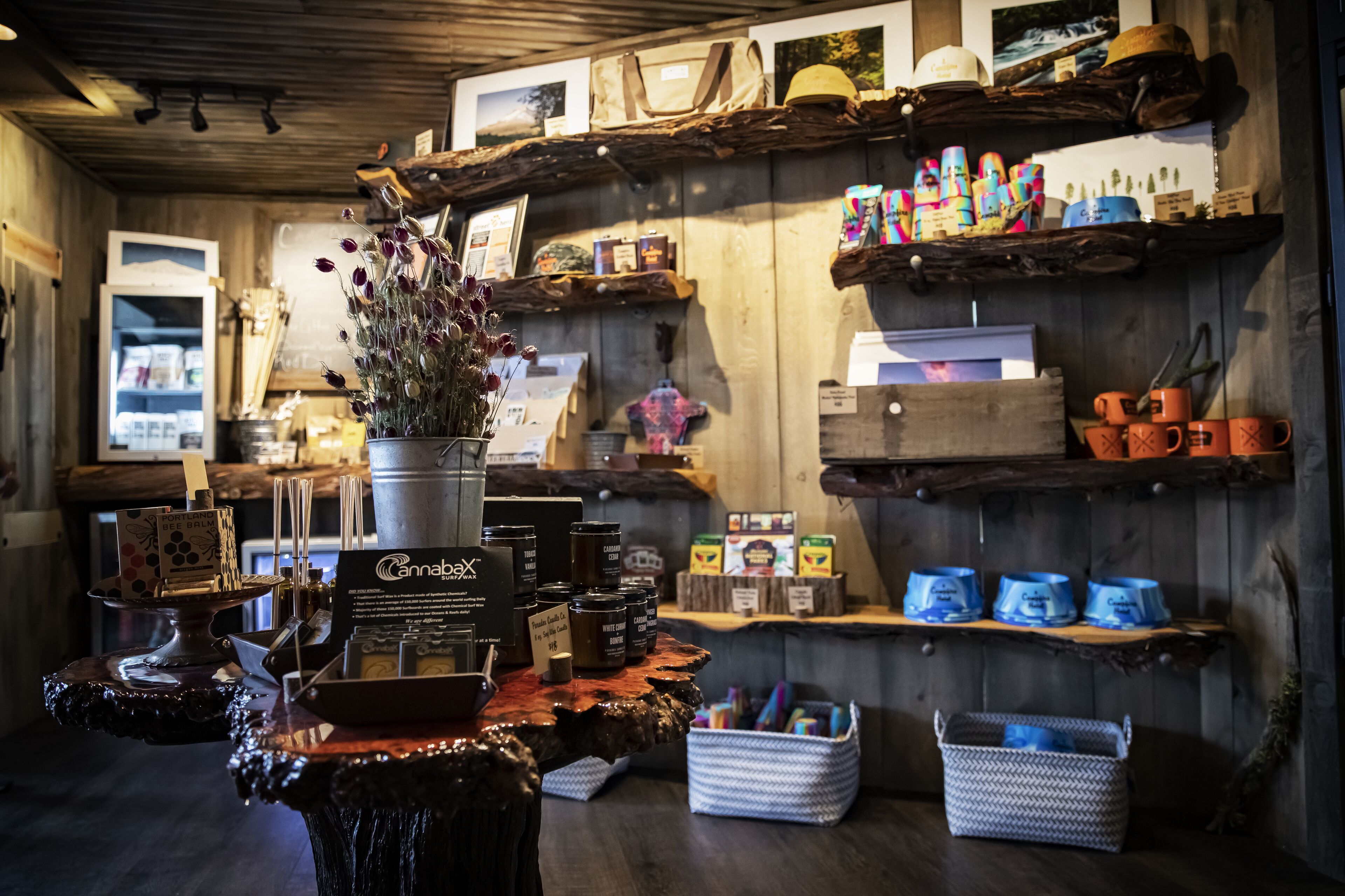 Een rustiek winkelinterieur met houten planken waarop diverse artikelen staan, een vaas met bloemen en een koelkast op de achtergrond.