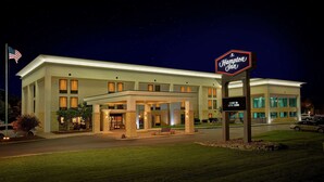 Un hotel Hampton Inn con una entrada bien iluminada, un área de espera cubierta y un letrero invitando a los huéspedes a entrar y disfrutar de la piscina.