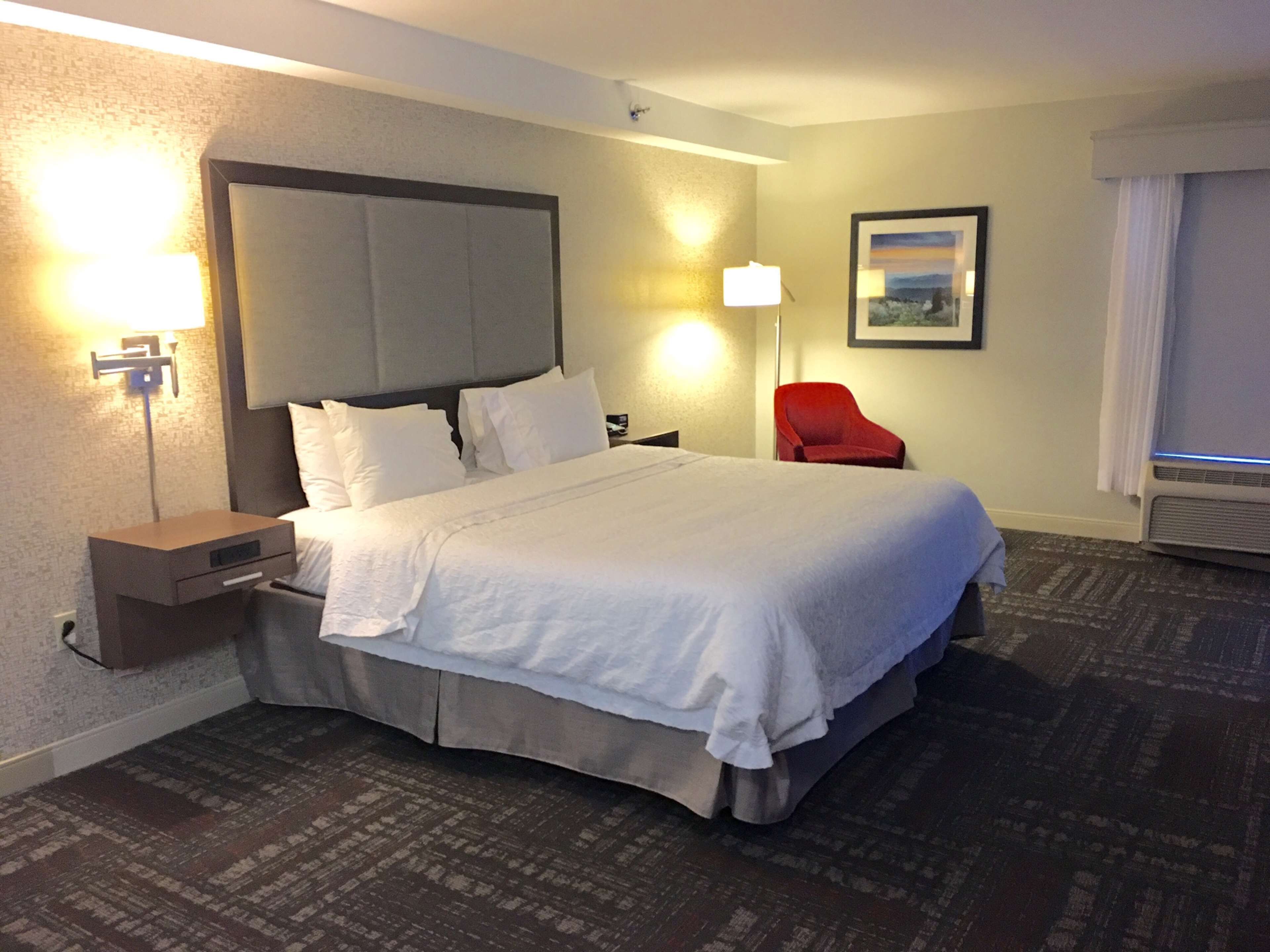 Habitación de hotel con una cama grande, una mesita de noche, una silla roja, una lámpara de pared y un cuadro enmarcado.