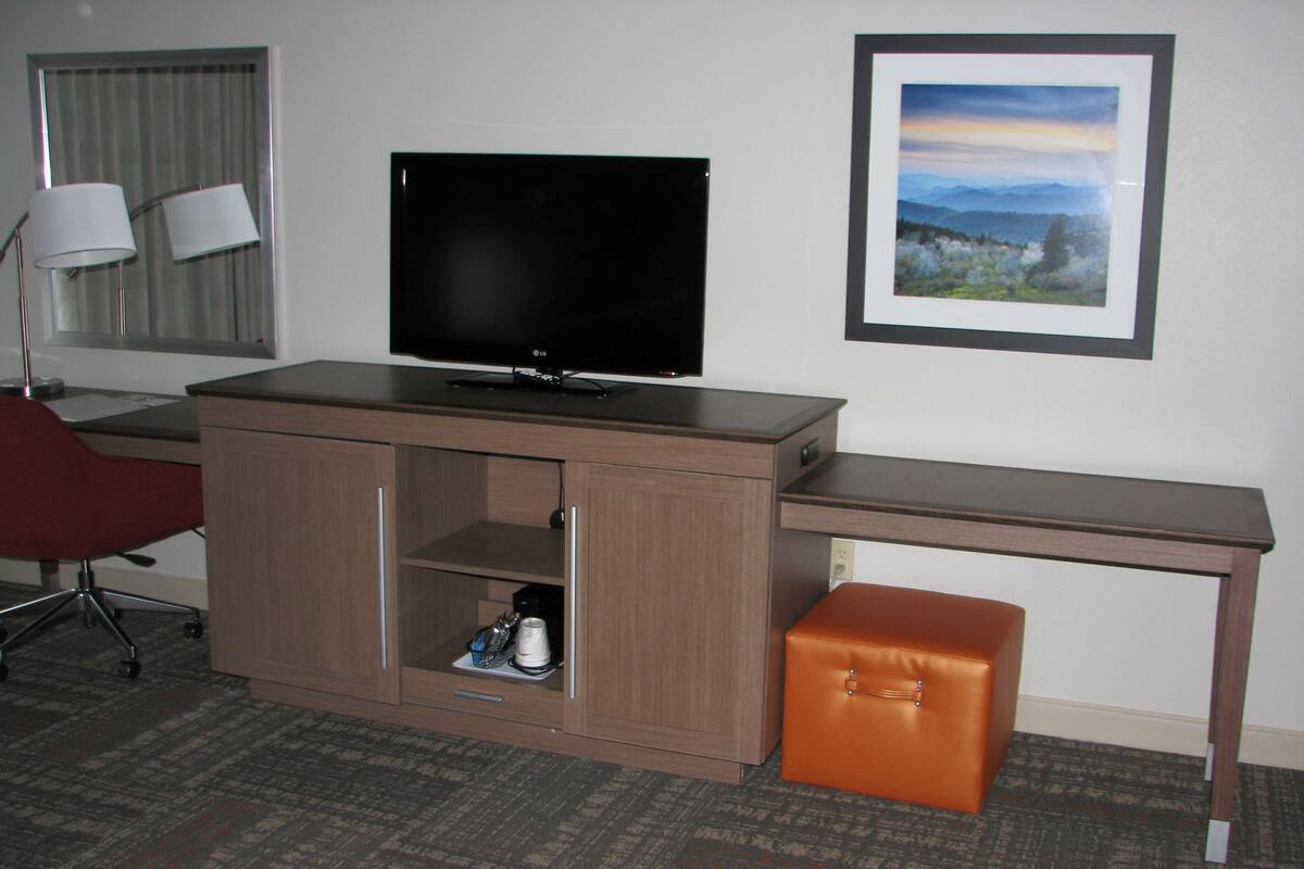 A hotel room with a wooden desk, a television, a red chair, a small cabinet with a shelf, and a framed picture on the wall.