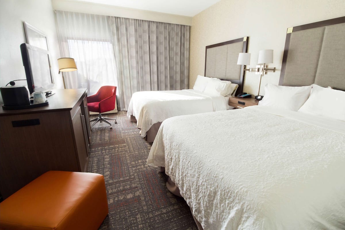 A hotel room with two beds, a desk with a computer, a red chair, and a small orange ottoman.