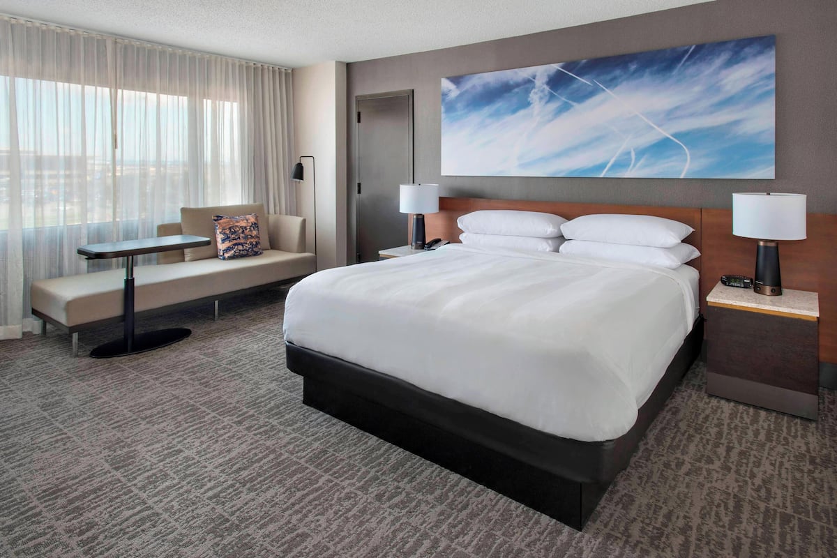 Habitación de hotel con una cama grande, un sofá, una mesita y un escritorio. Hay un cuadro en la pared y una ventana con cortinas.