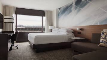 Habitación de hotel con una cama grande, un escritorio, una silla y vista a la ciudad a través de la ventana.