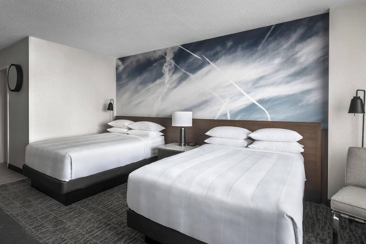 Una habitación de hotel con dos camas, un cuadro abstracto grande en la pared, un espejo redondo, un escritorio con una lámpara y una silla.