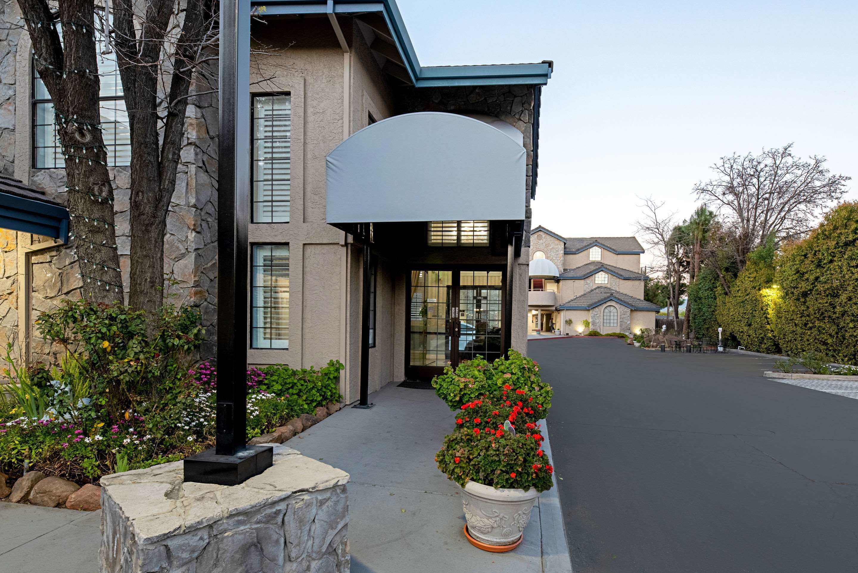 A modern building with a stone facade, a covered entrance, and a well-maintained garden with flowering plants.