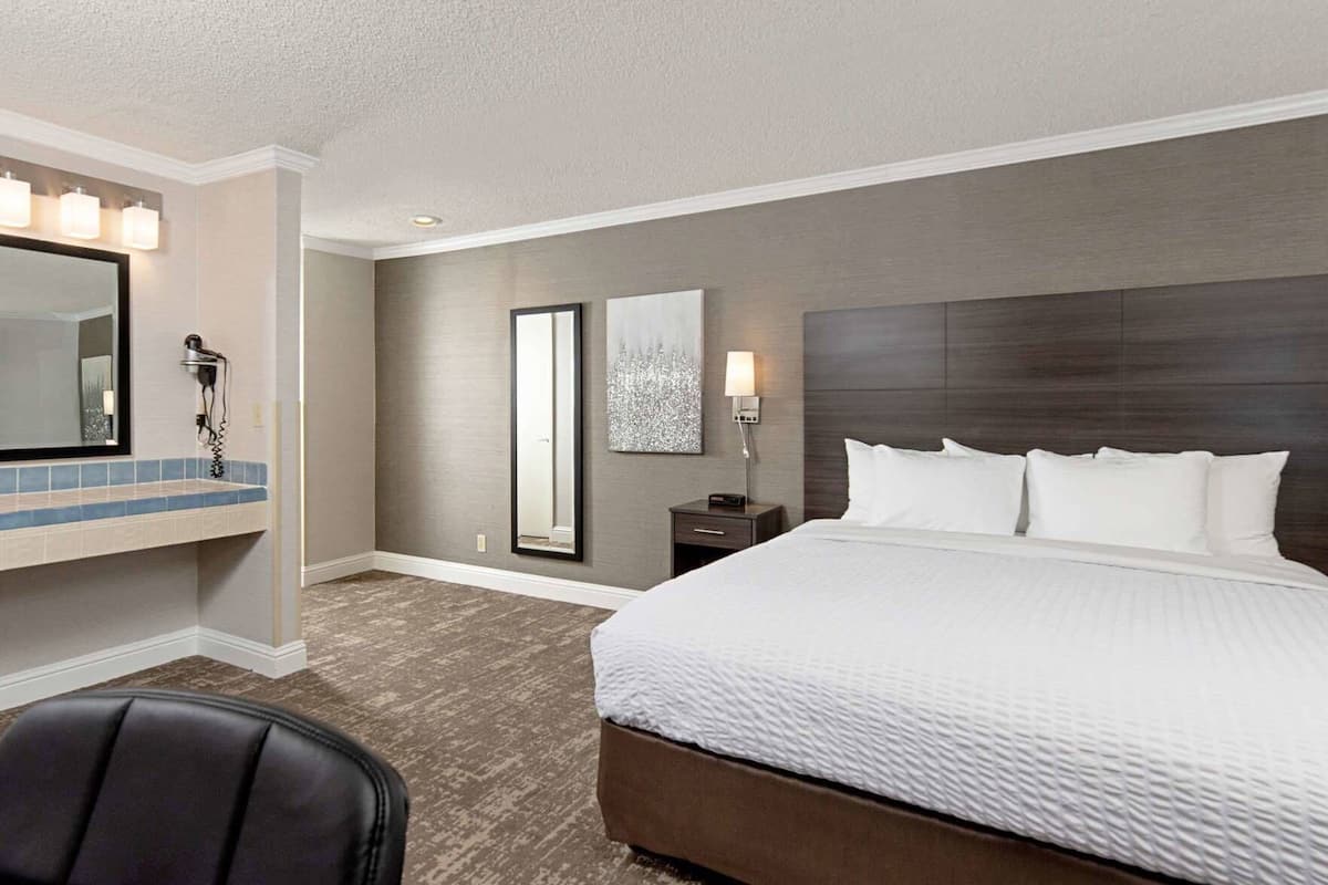A hotel room with a large bed, a desk with a chair, a mirror, and a nightstand with a lamp.
