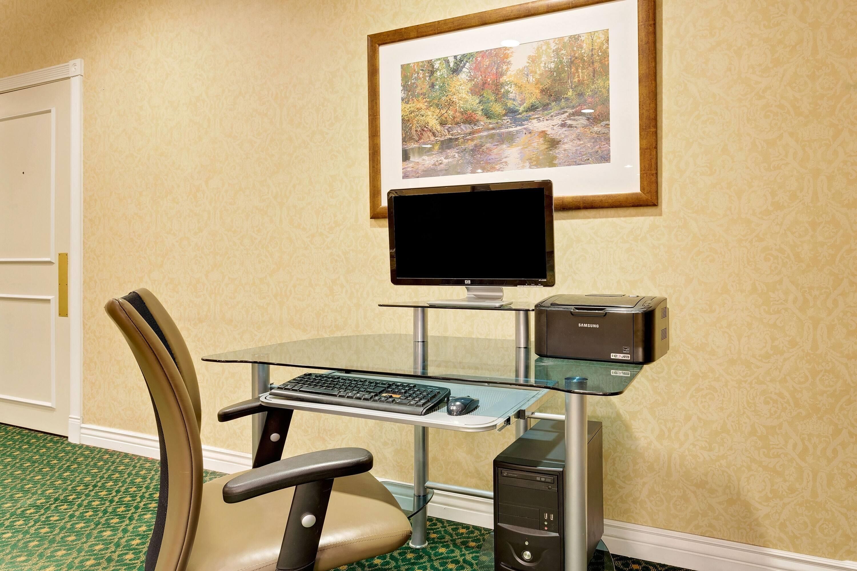 A hotel room with a glass desk, a computer, a chair, and a framed picture on the wall.