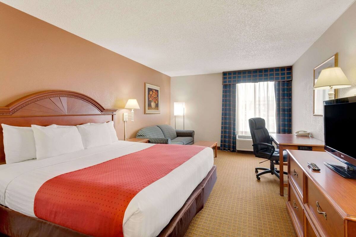 Habitación de hotel con una cama grande, un escritorio con silla, televisión y ventana con cortinas.
