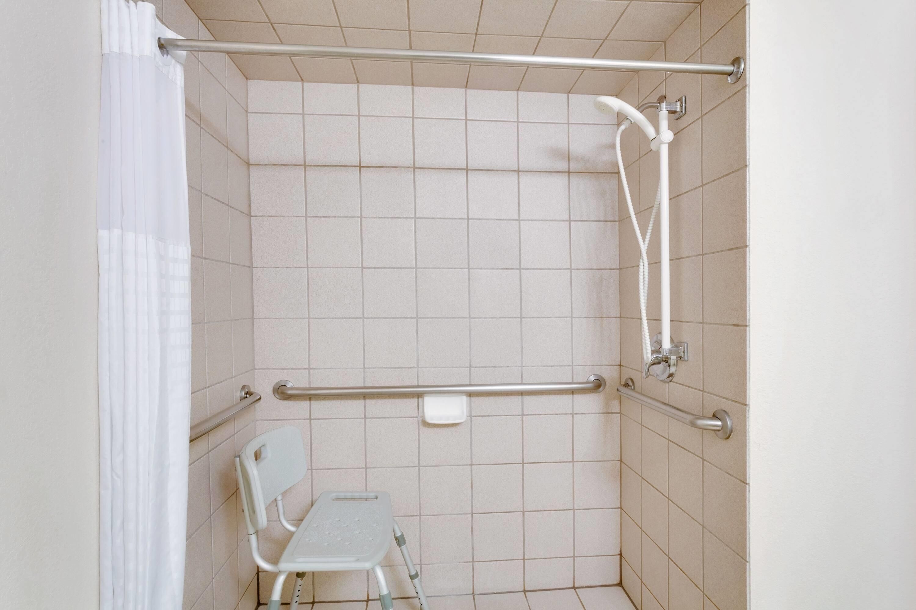 A shower area with a grab bar, a white chair, and a handheld showerhead.