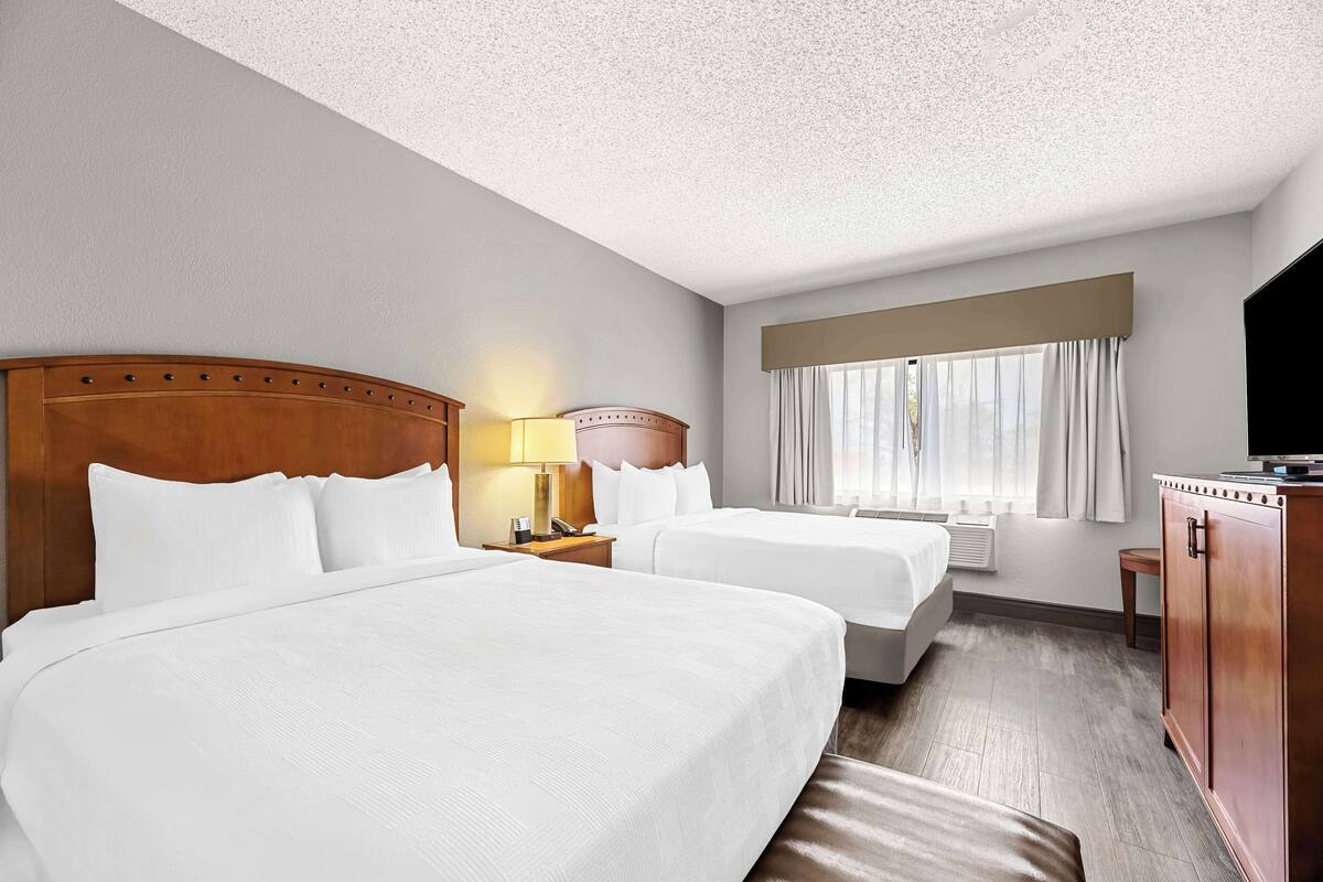 A hotel room with two beds, a wooden headboard, a nightstand with a lamp, a TV, and a window with curtains.