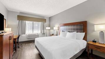 Una habitación de hotel con una cama grande, un escritorio, una silla, un televisor, una ventana con cortinas y una mesita de noche con lámpara.