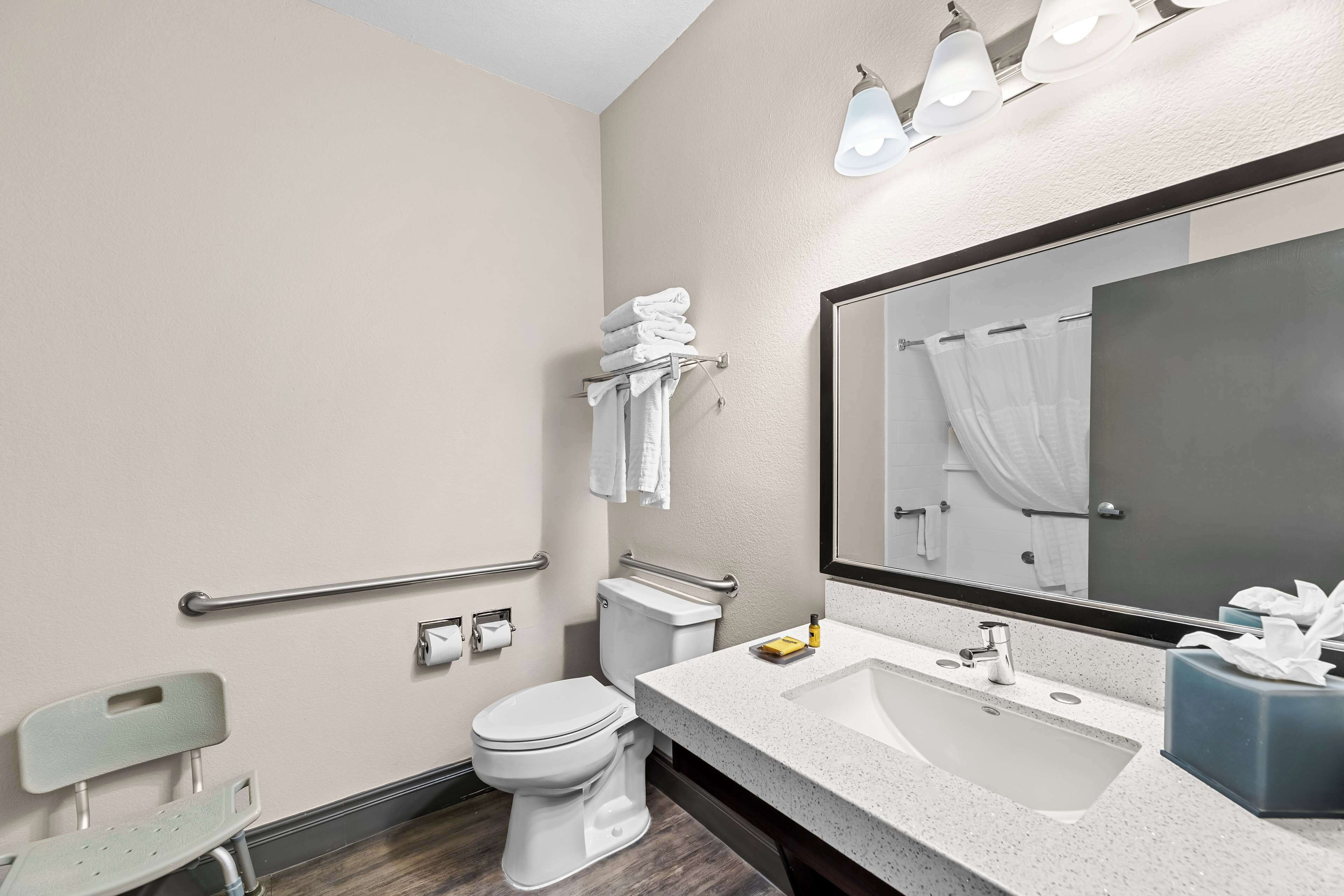 Et moderne badeværelse med toilet, vask og spejl. Der er gelændere og en bænk.
