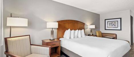 A hotel room with a large bed, two bedside tables with lamps, a wooden headboard, a chair, and a framed picture on the wall.