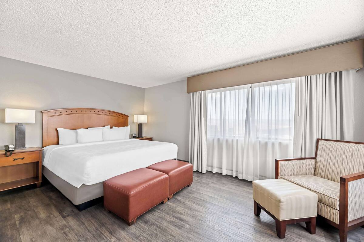 A hotel room with a large bed, a wooden headboard, a nightstand with a lamp, a chair, and a window with curtains.