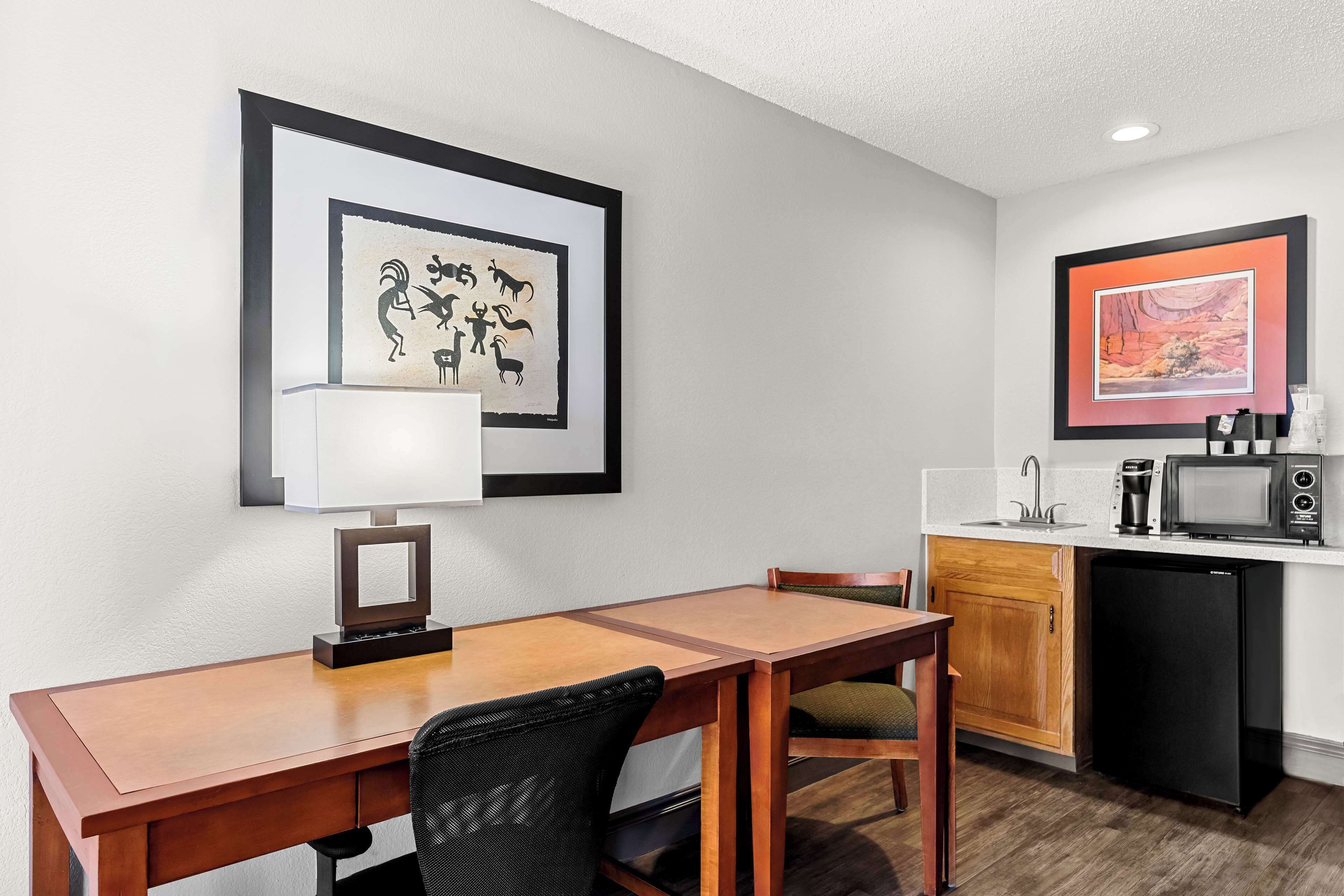 Een compacte hotelkamer met een houten eettafel, een zwarte bureaustoel, een kleine kitchenette met een magnetron en een koelkast. Er hangen ingelijste foto’s aan de muur.