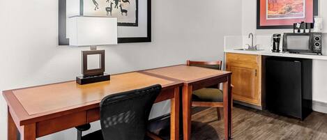 Een compacte hotelkamer met een houten eettafel, een zwarte bureaustoel, een kleine kitchenette met een magnetron en een koelkast. Er hangen ingelijste foto’s aan de muur.