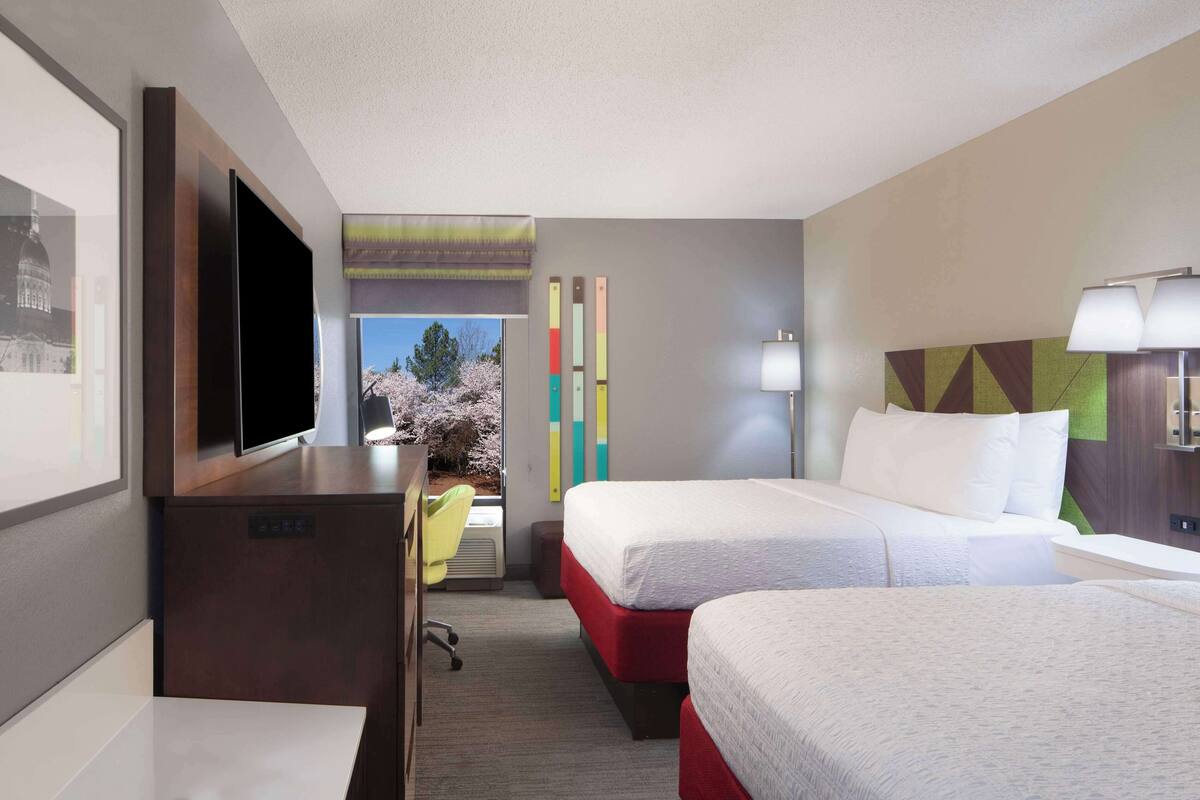 A hotel room with two beds, a desk, a TV, and a window with a view of trees.