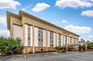Un hotel Hampton Inn con una fachada moderna, un estacionamiento y un cielo azul despejado.
