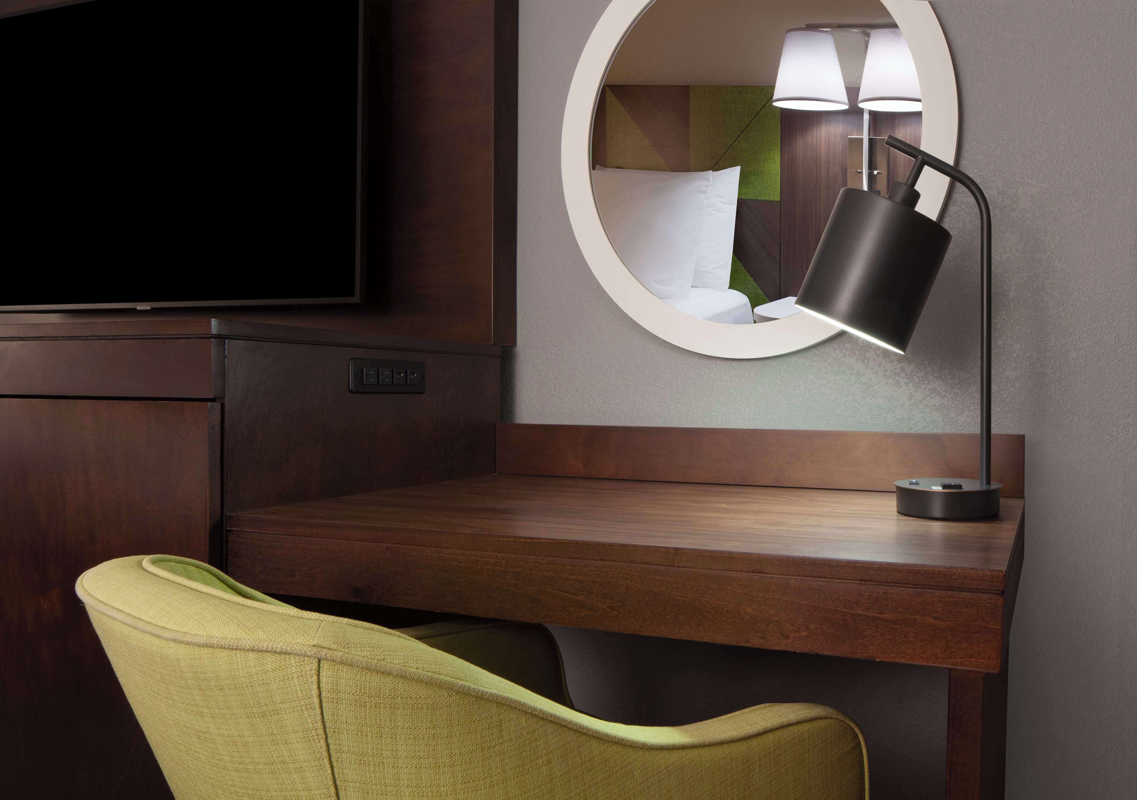 A hotel room with a wooden desk, a green chair, a round mirror, and a flat-screen TV.