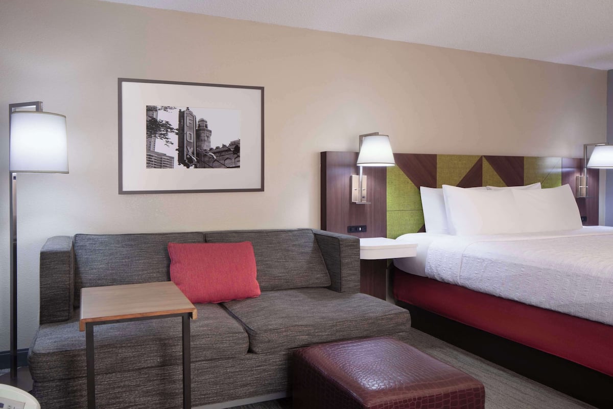 A hotel room with a grey sofa, a small table, a red pillow, a bed with white bedding, and a framed picture on the wall.