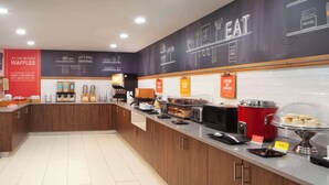 Una cafetería moderna con una pared oscura que presenta escritura al estilo de pizarra, una máquina de café, un microondas y una exhibición de gofres y galletas.