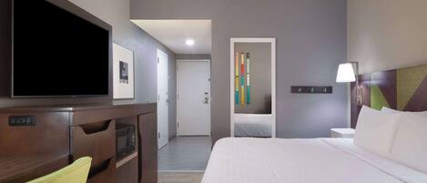 A hotel room with a bed, a TV, a desk with a computer, a chair, a lamp, and a colorful wall decoration.