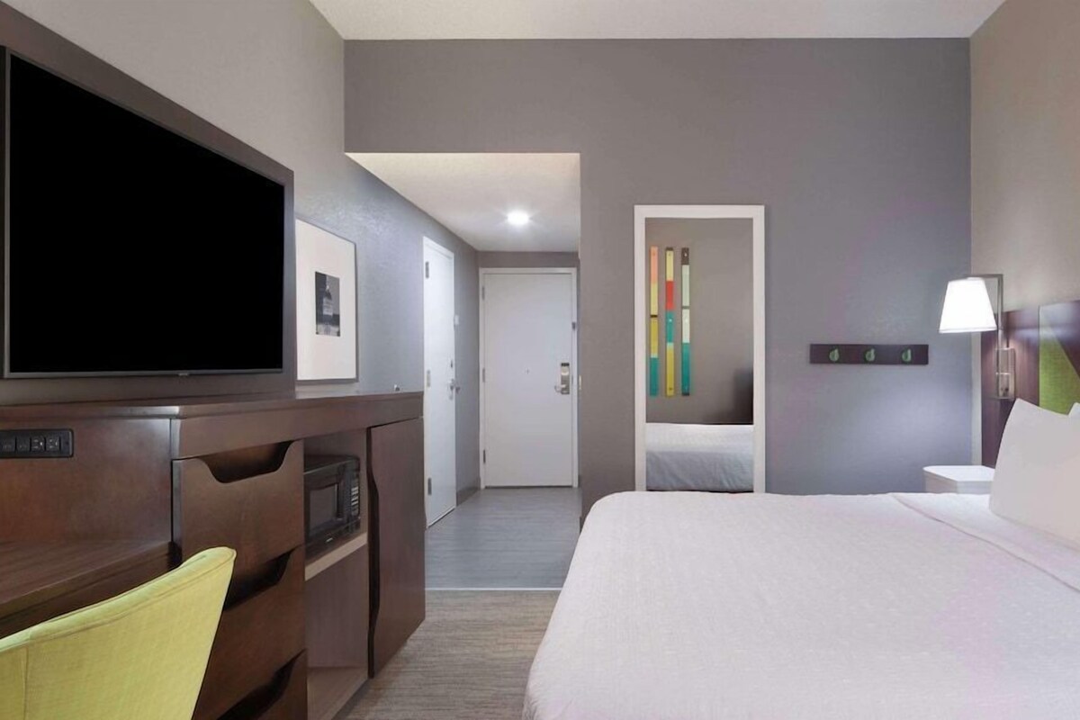 A hotel room with a bed, a TV, a desk, a chair, a door, and a wall with a colorful artwork.