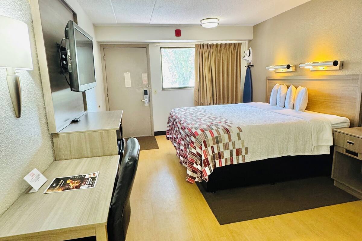 Habitación de hotel con cama, escritorio, silla, televisor y ventana con cortinas.