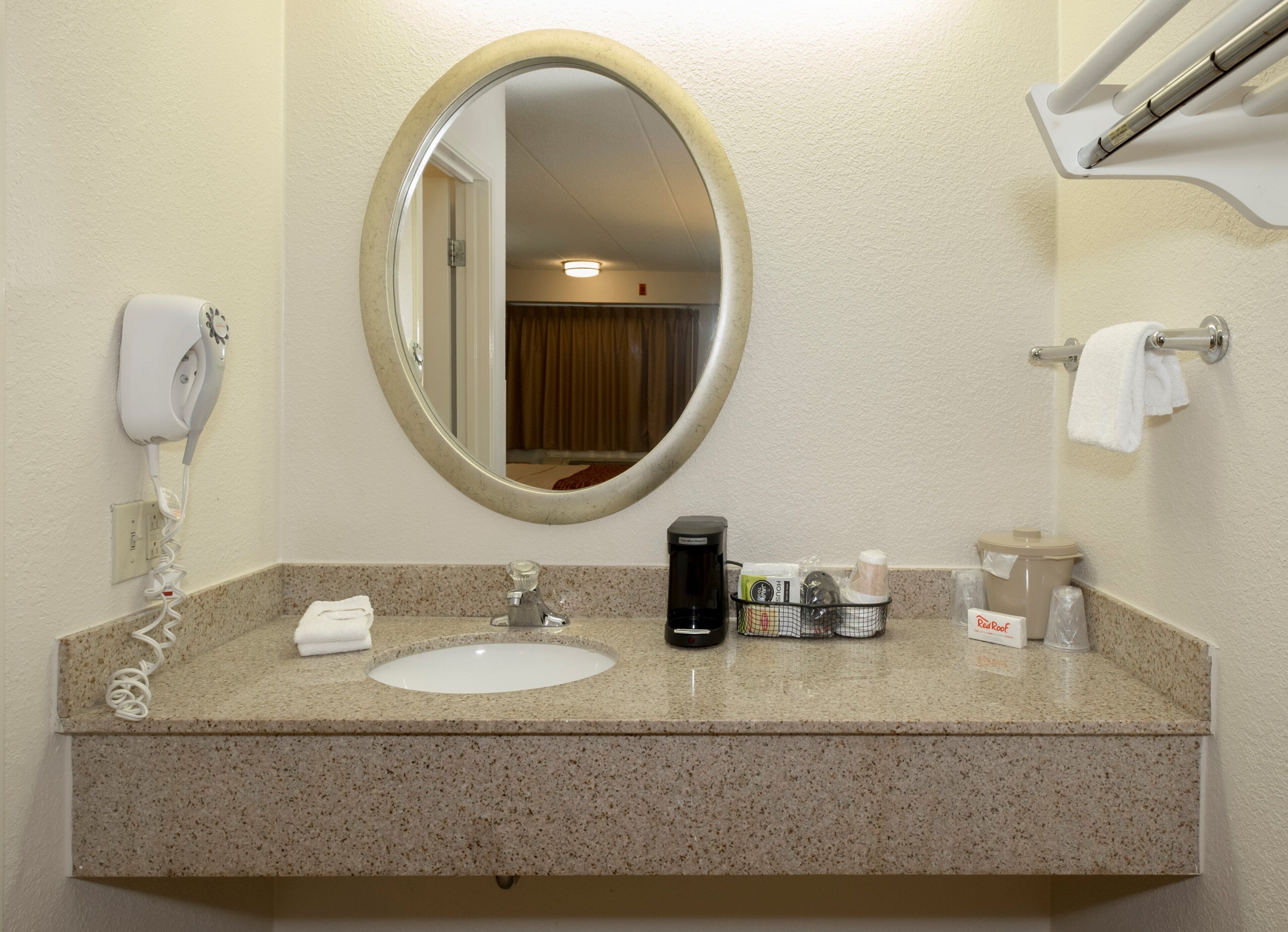 A bathroom with a round mirror, a sink, a hair dryer, a coffee maker, and toiletries.