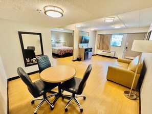 Habitación de hotel con una mesa redonda, dos sillas negras, un sofá, un televisor, un minibar y un espejo.