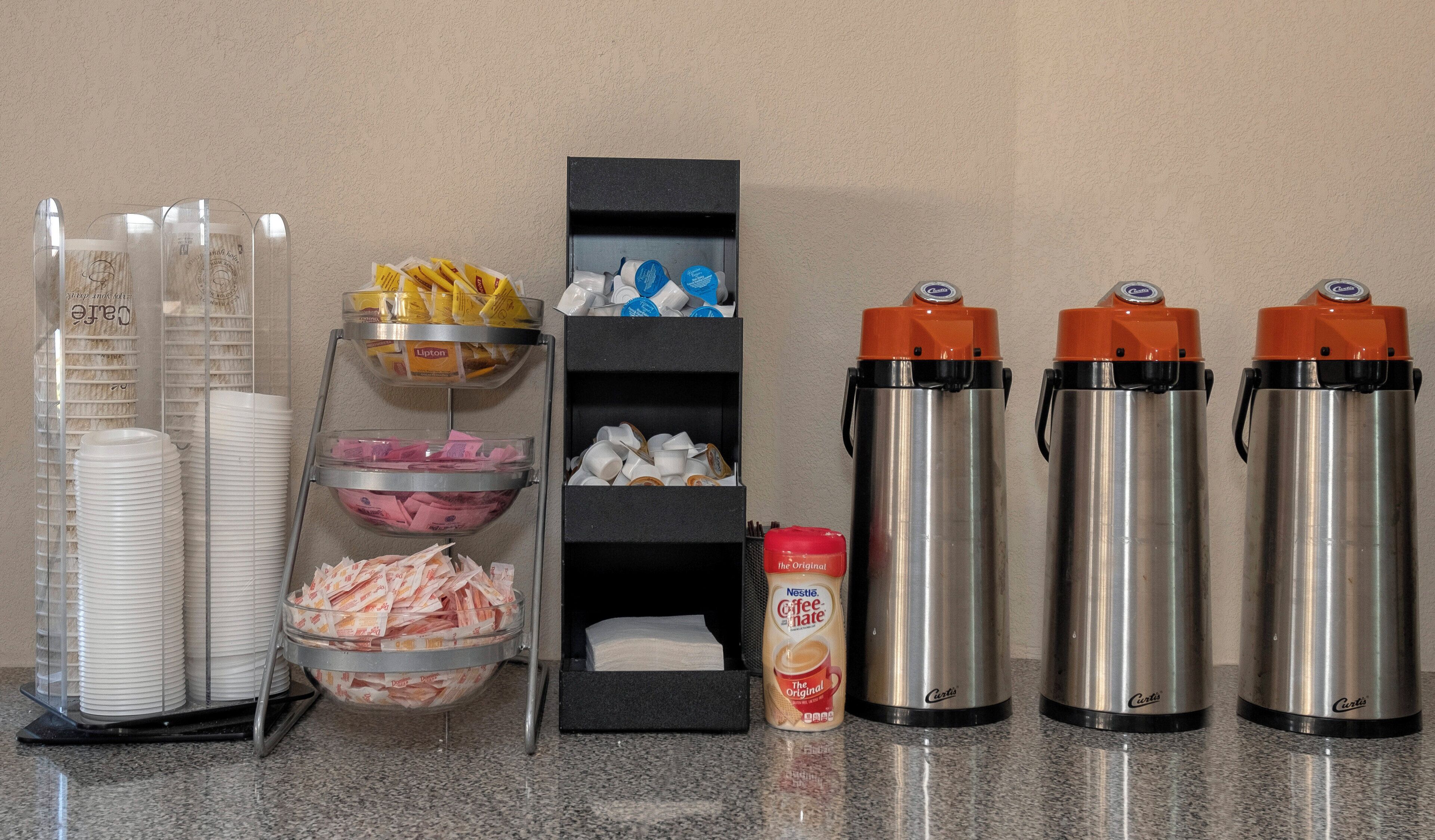 A buffet setup with clear glass containers for food, a coffee station with disposable cups, and two stainless steel thermos flasks.