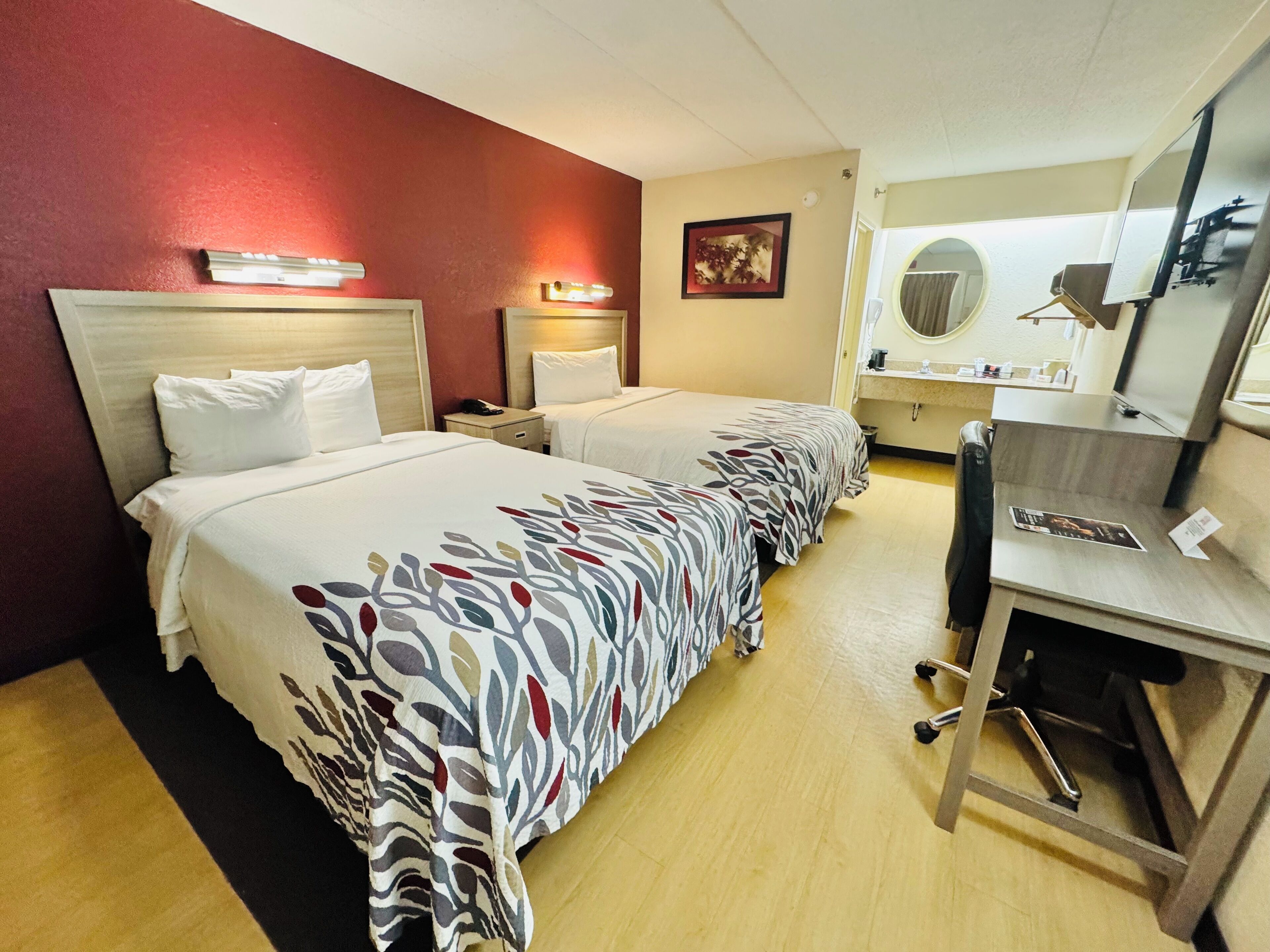 A hotel room with two beds, a desk, a chair, a mirror, and a small bathroom area.