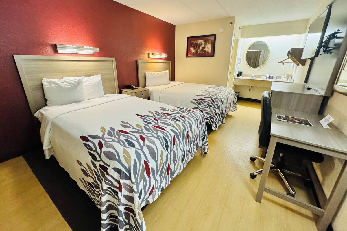 Ein Hotelzimmer mit zwei Betten, einem Schreibtisch, einem Stuhl, einem Spiegel und einem kleinen Badebereich.