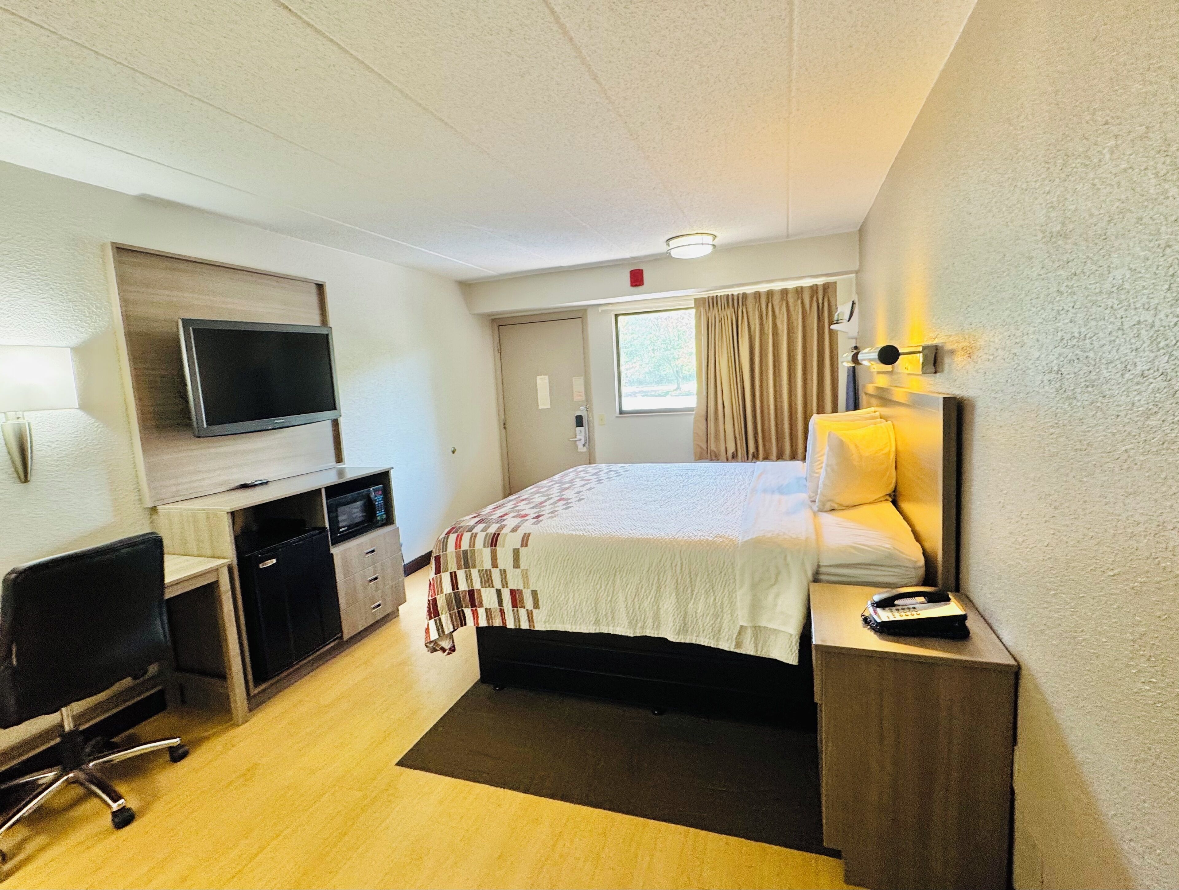 A hotel room with a bed, a desk with a computer, a TV, and a window with curtains.