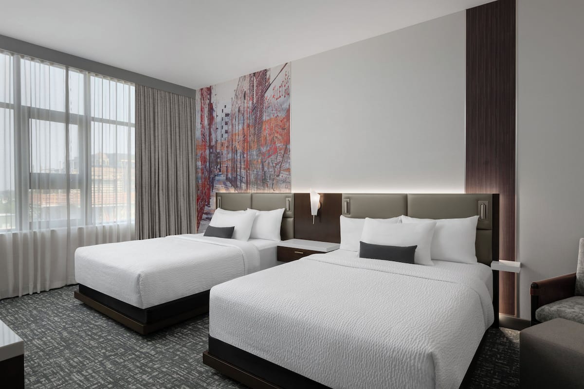 A hotel room with two beds, a large window with curtains, a painting on the wall, and a nightstand with a lamp.