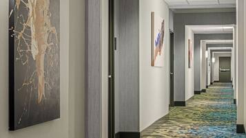 A hotel hallway with a patterned carpet, framed artwork on the walls, and doors leading to rooms.