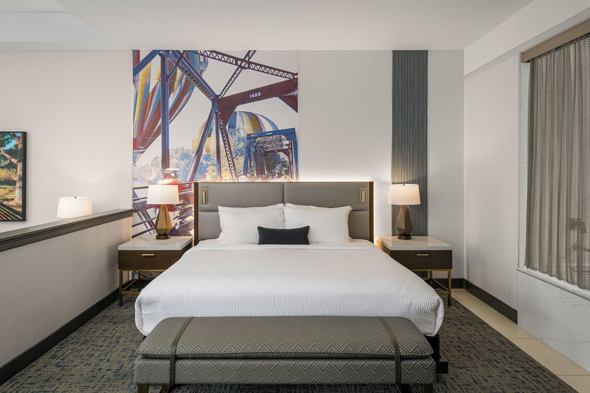 A hotel room with a large bed, two bedside tables, a bench, a wall mural, and a window with blinds.