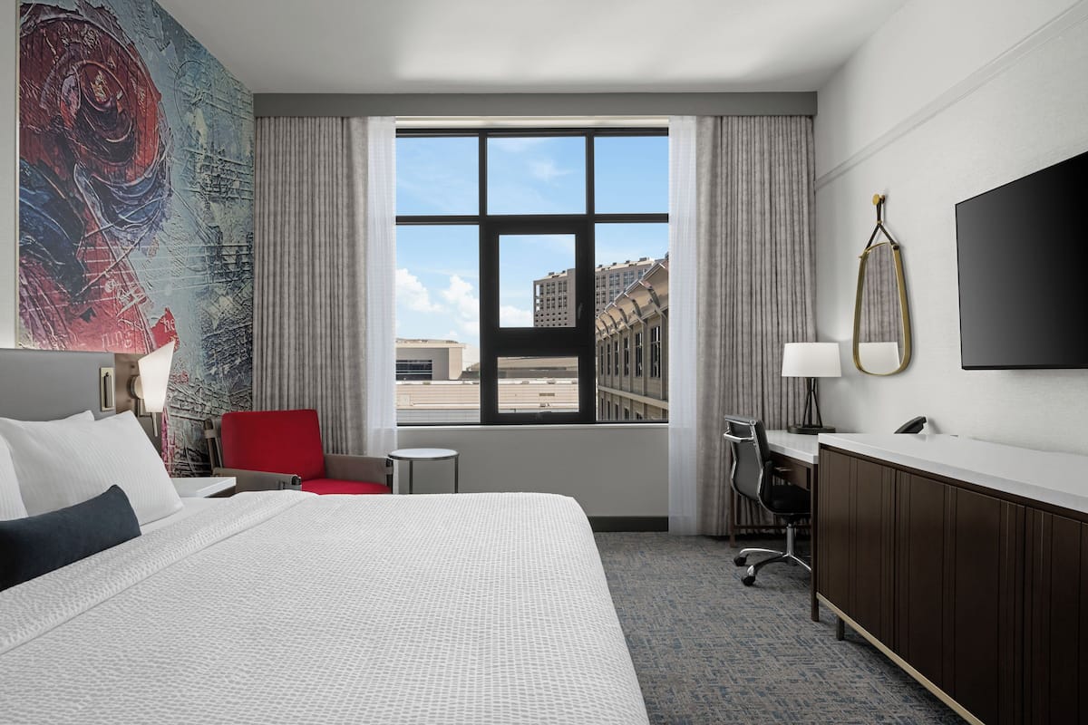 A hotel room with a large bed, a desk with a chair, a TV, and a window with a city view.