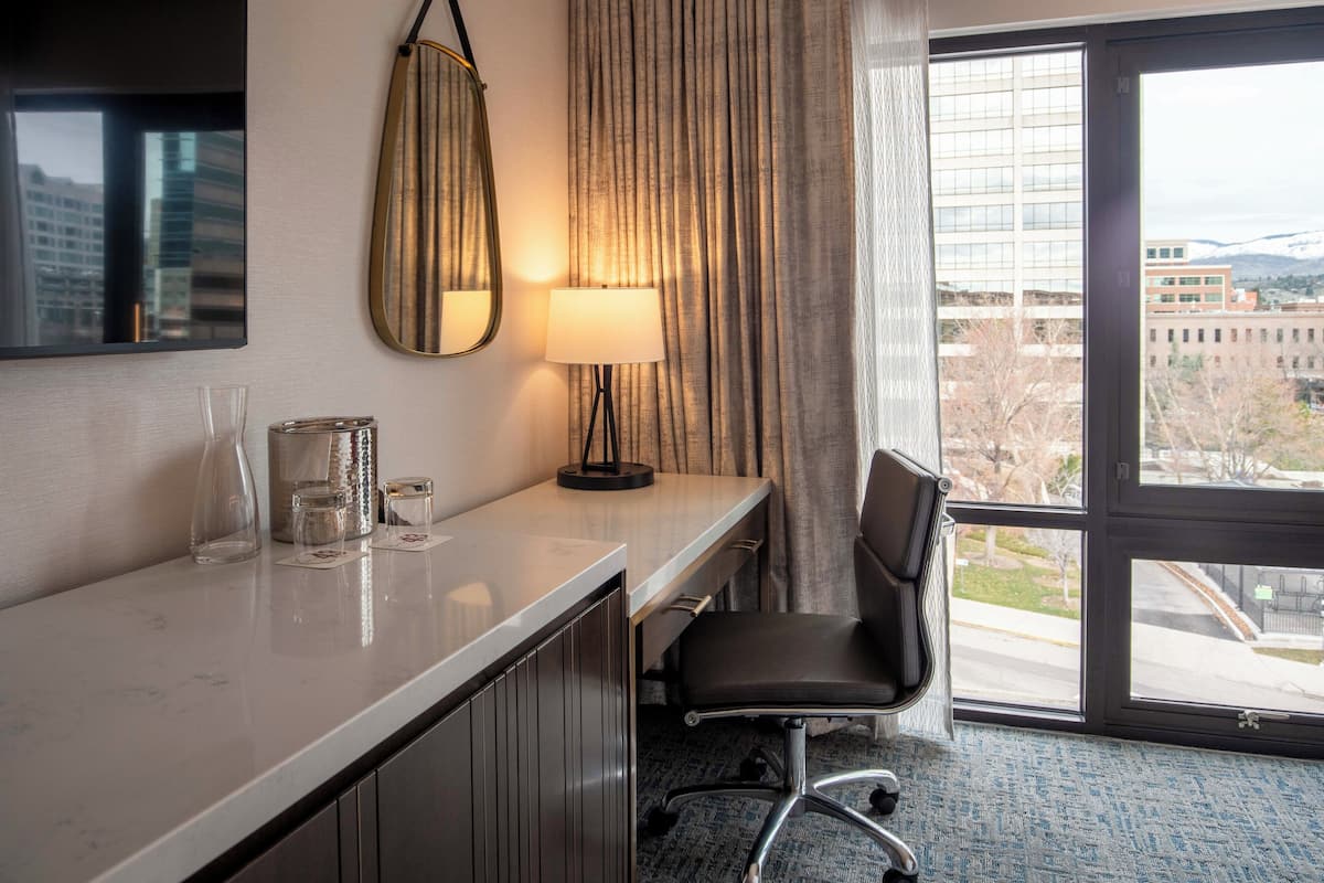 A hotel room with a desk, chair, lamp, and large window with a view of buildings and mountains.