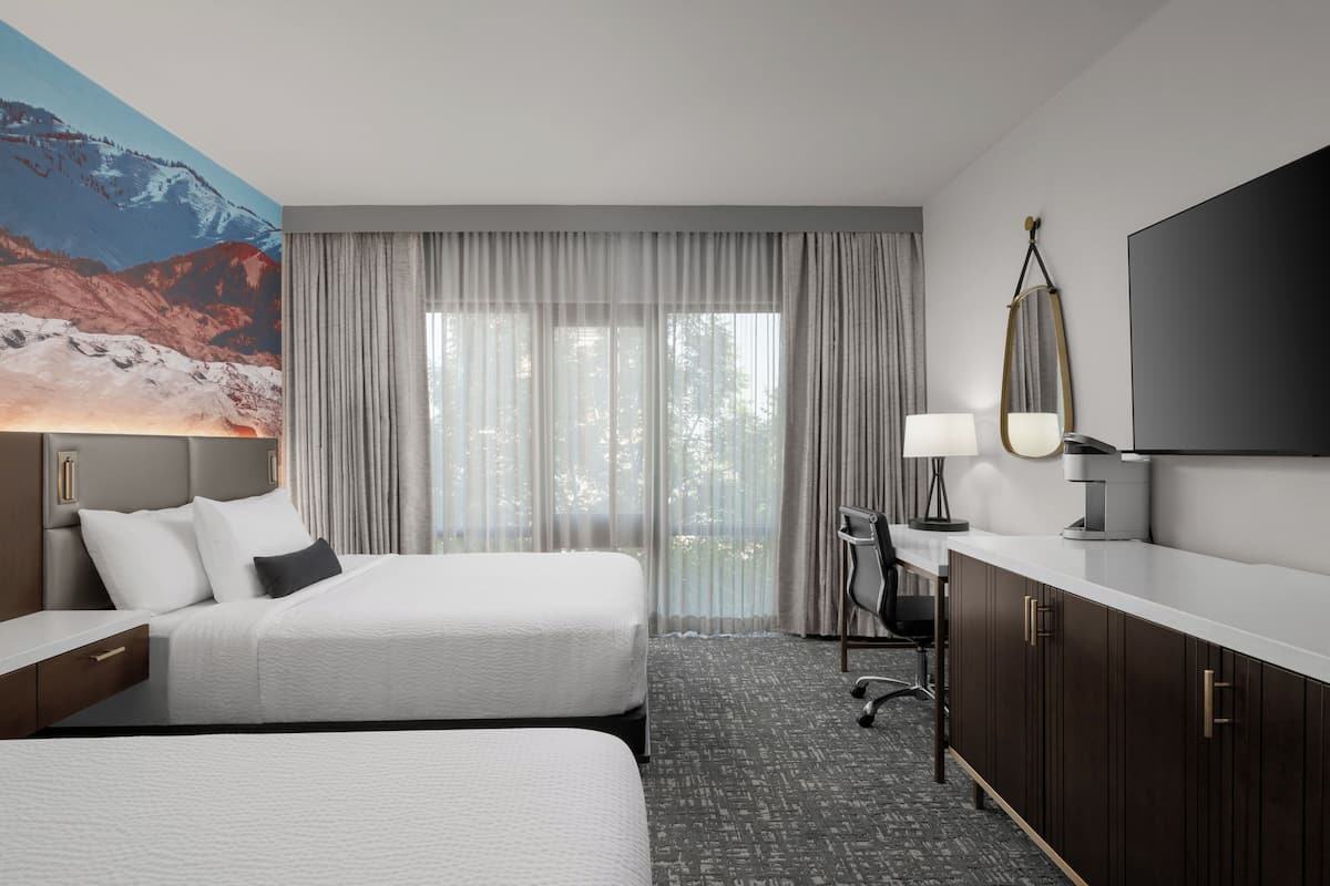 A hotel room with two beds, a desk, a chair, a TV, and a large window with curtains.