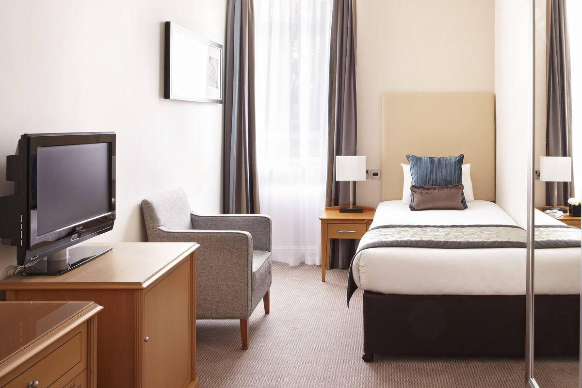 A hotel room with a bed, a desk with a TV, a chair, a bedside table with a lamp, and a large mirror.
