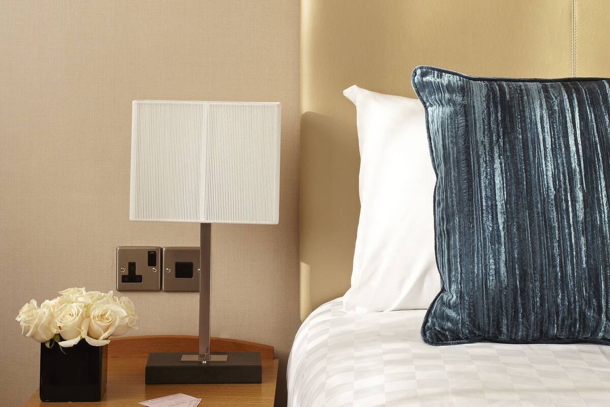 A neatly made bed with a headboard, a bedside table with a lamp, a vase of flowers, and a nightstand with a drawer.