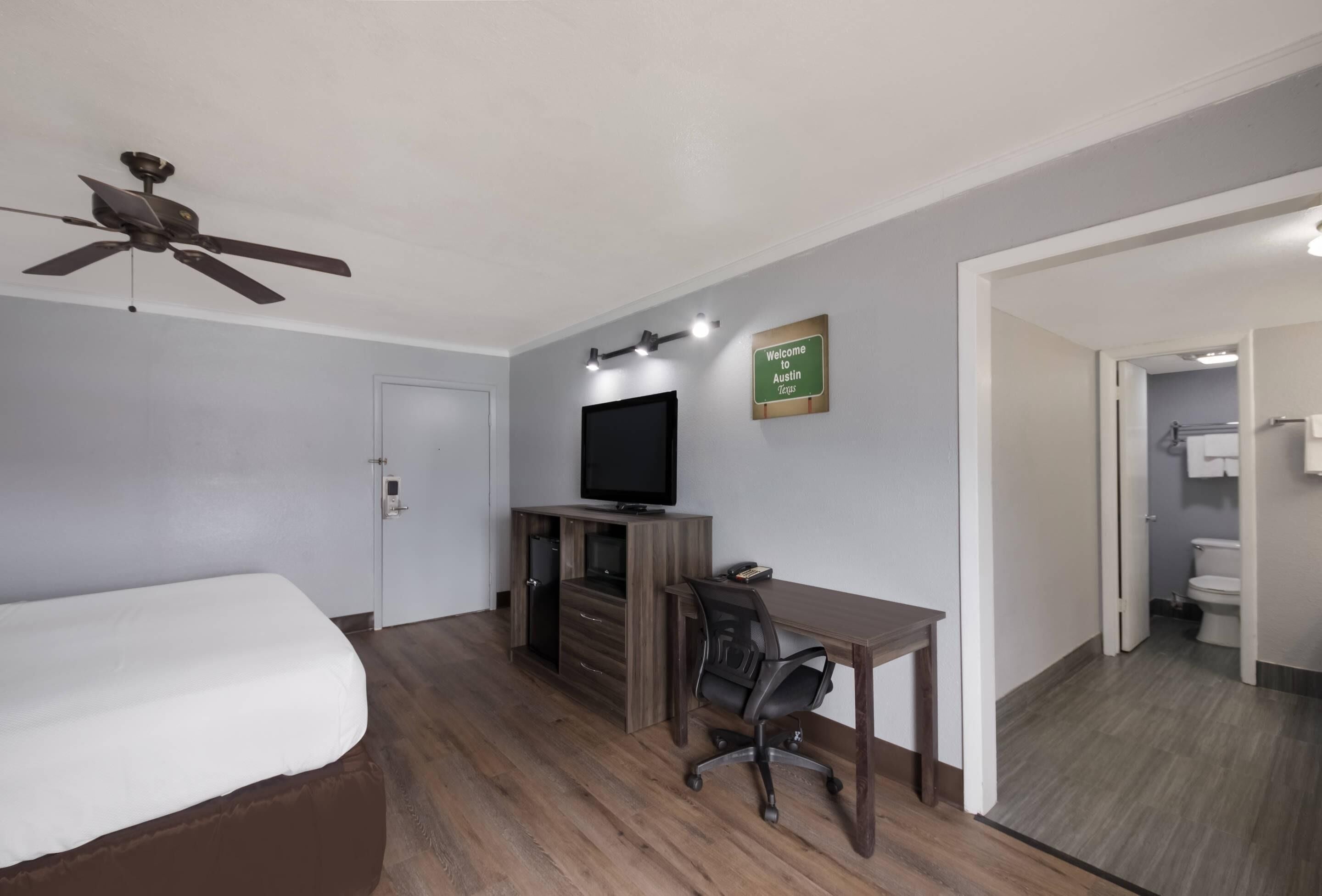 A bedroom with a bed, a desk, a chair, a ceiling fan, a television, and a sign that says "Welcome Austin Texas".