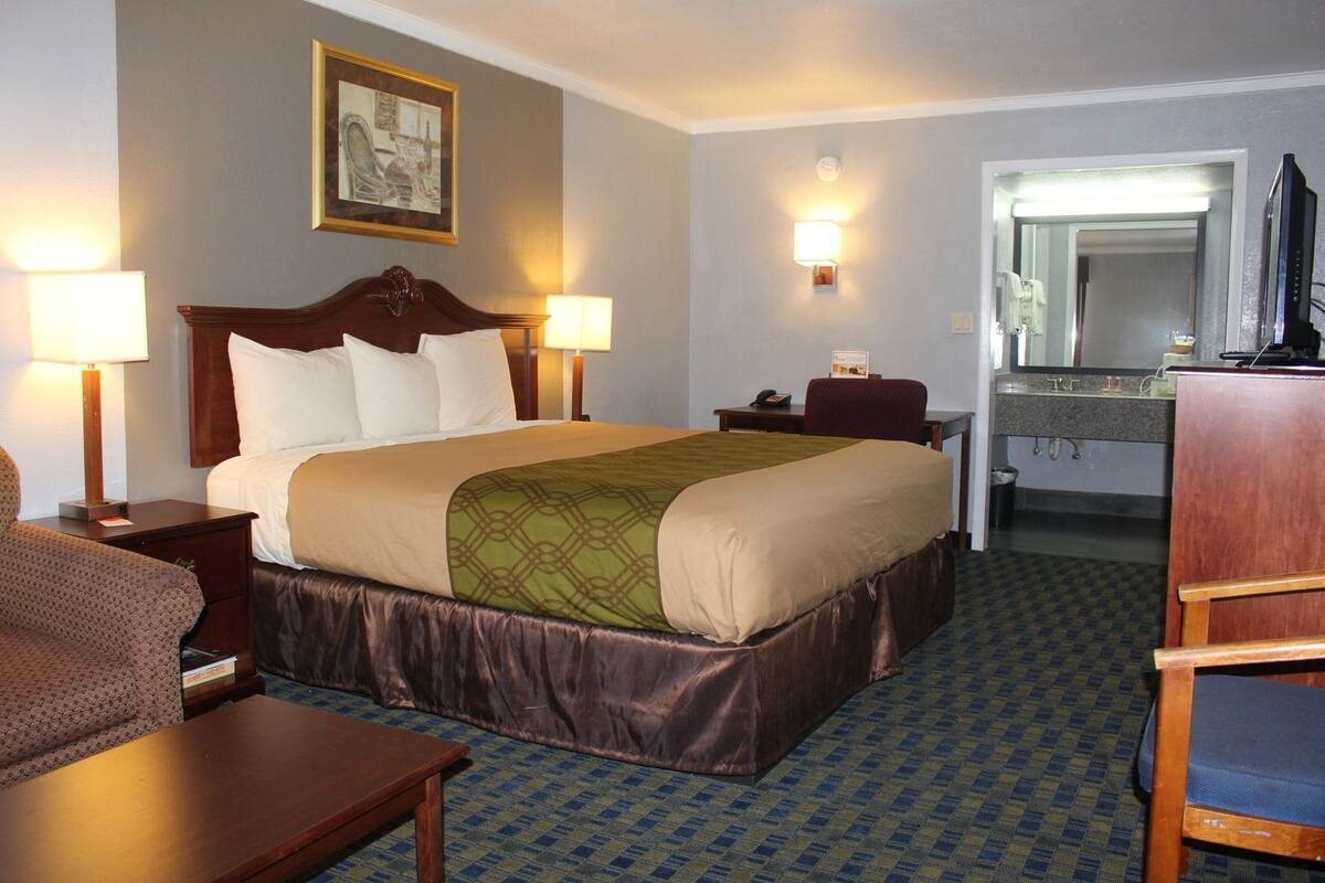 A hotel room with a bed, two bedside lamps, a desk, a chair, a mirror, and a bathroom visible through an open door.