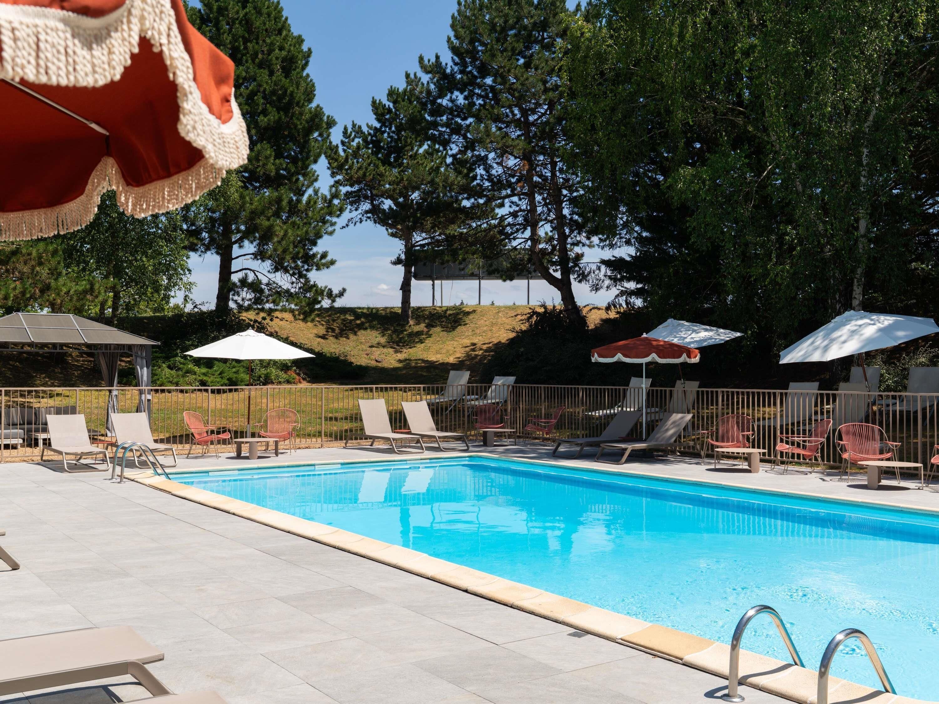 A swimming pool with lounge chairs and umbrellas, surrounded by trees and a hilly landscape.