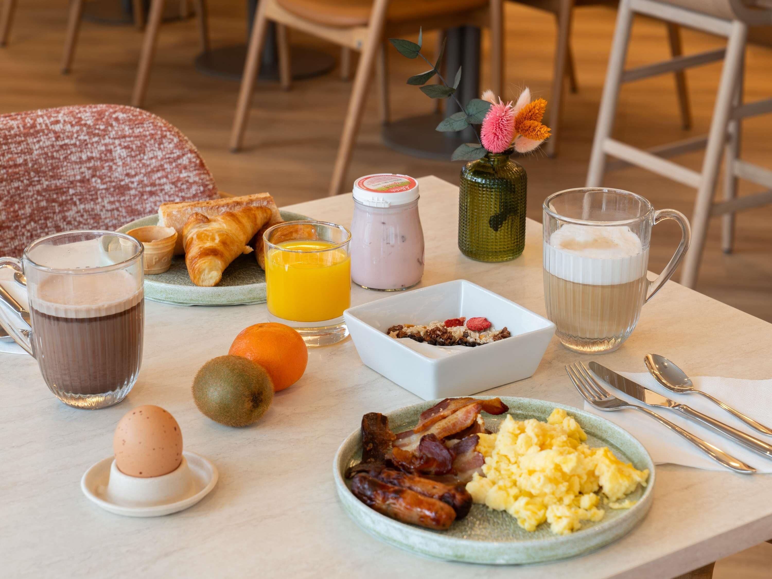 A breakfast table with croissants, orange juice, coffee, scrambled eggs, bacon, and a small vase with flowers.