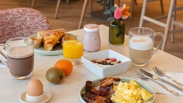 A breakfast table with croissants, orange juice, coffee, scrambled eggs, bacon, and a small vase with flowers.
